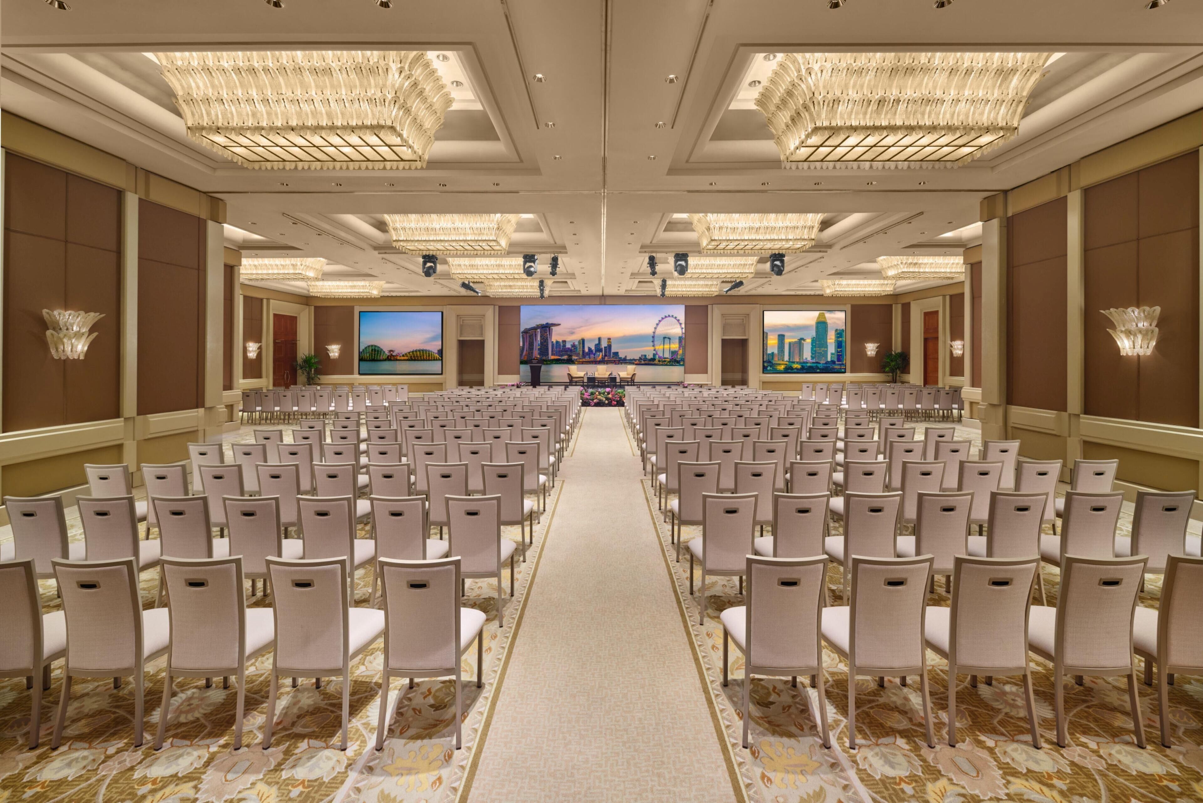 A conference room with rows of chairs, a carpeted floor, chandeliers, and a large screen displaying a cityscape.