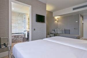 Ett hotellrum med en stor säng, en sittgrupp med bord och stolar, en platt-TV som är monterad på väggen och ett badrum med dusch och handfat.