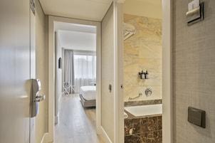 A hotel room with a modern design, featuring a large bed, a bathroom with a bathtub, and a hallway leading to another room.