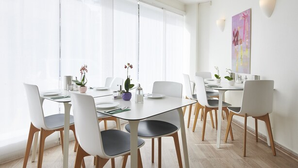 A modern dining area with a glass partition, white chairs, and a table set for six. - BALEGRA City Hotel Basel Contactless Self Check-in (Basel)