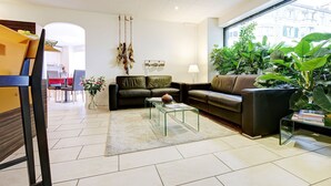 A modern living room with a sofa, coffee table, and a dining area in the background. - BALEGRA City Hotel Basel Contactless Self Check-in (Basel)