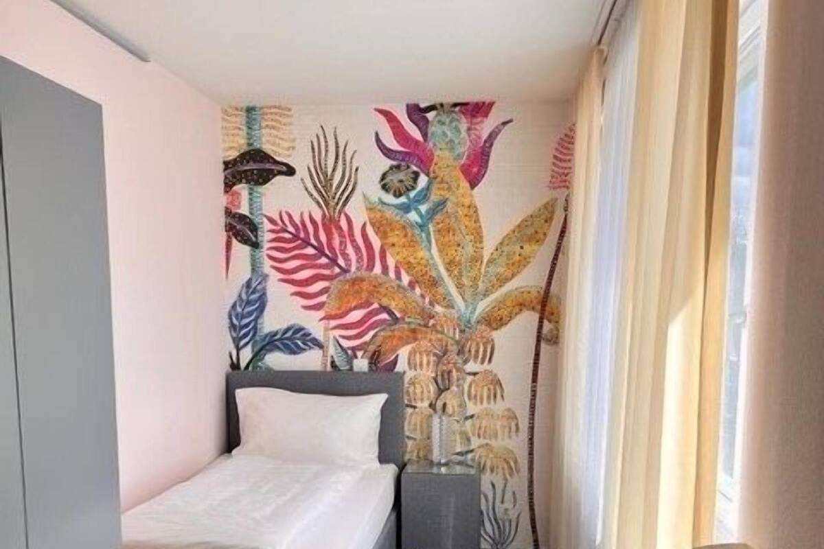 A compact hotel room with a bed, a small table, a chair, and a wall with a colorful tropical mural.