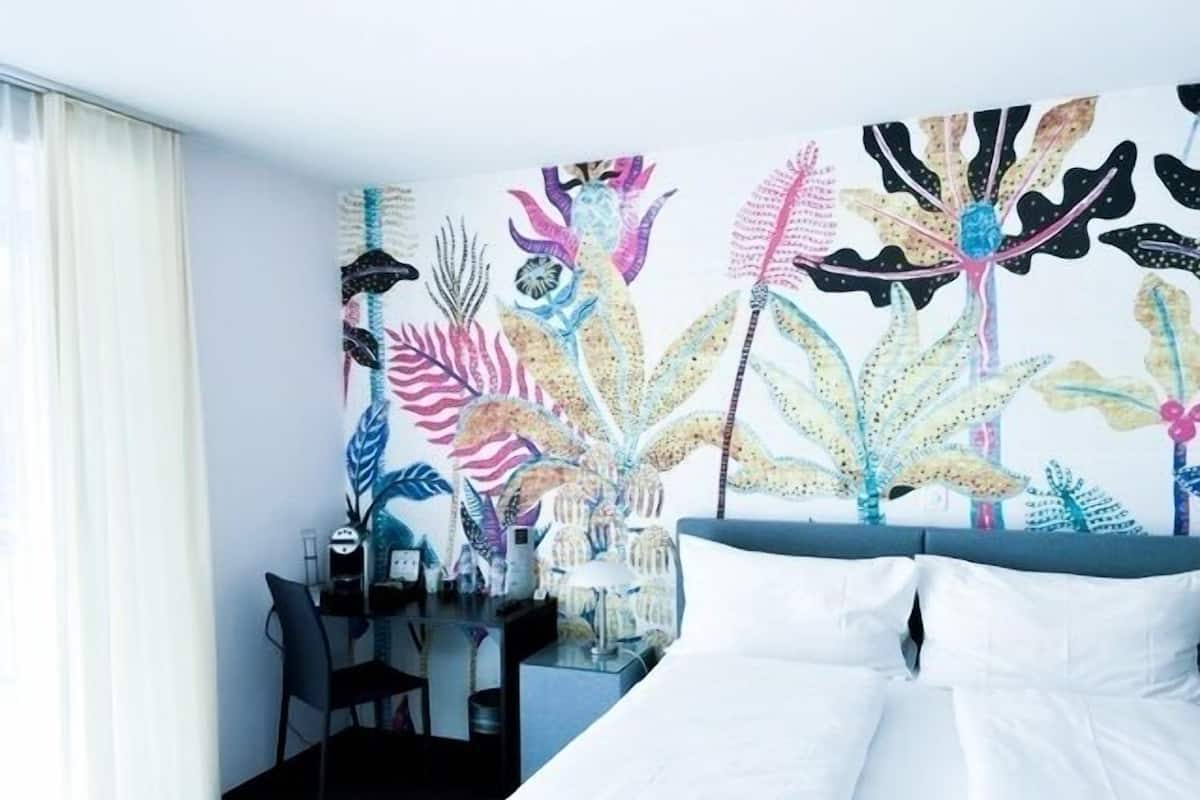 A hotel room with a bed, a desk, and a chair. There is a large, colorful wall mural with various plant designs.