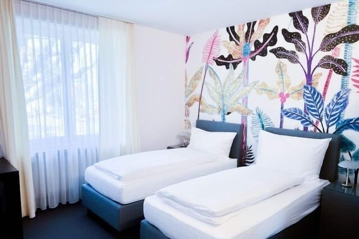A hotel room with two single beds, a large window with sheer curtains, and a colorful wall mural.
