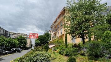 A hotel with a sign reading "HOTEL ELITE" surrounded by lush greenery and parked cars.