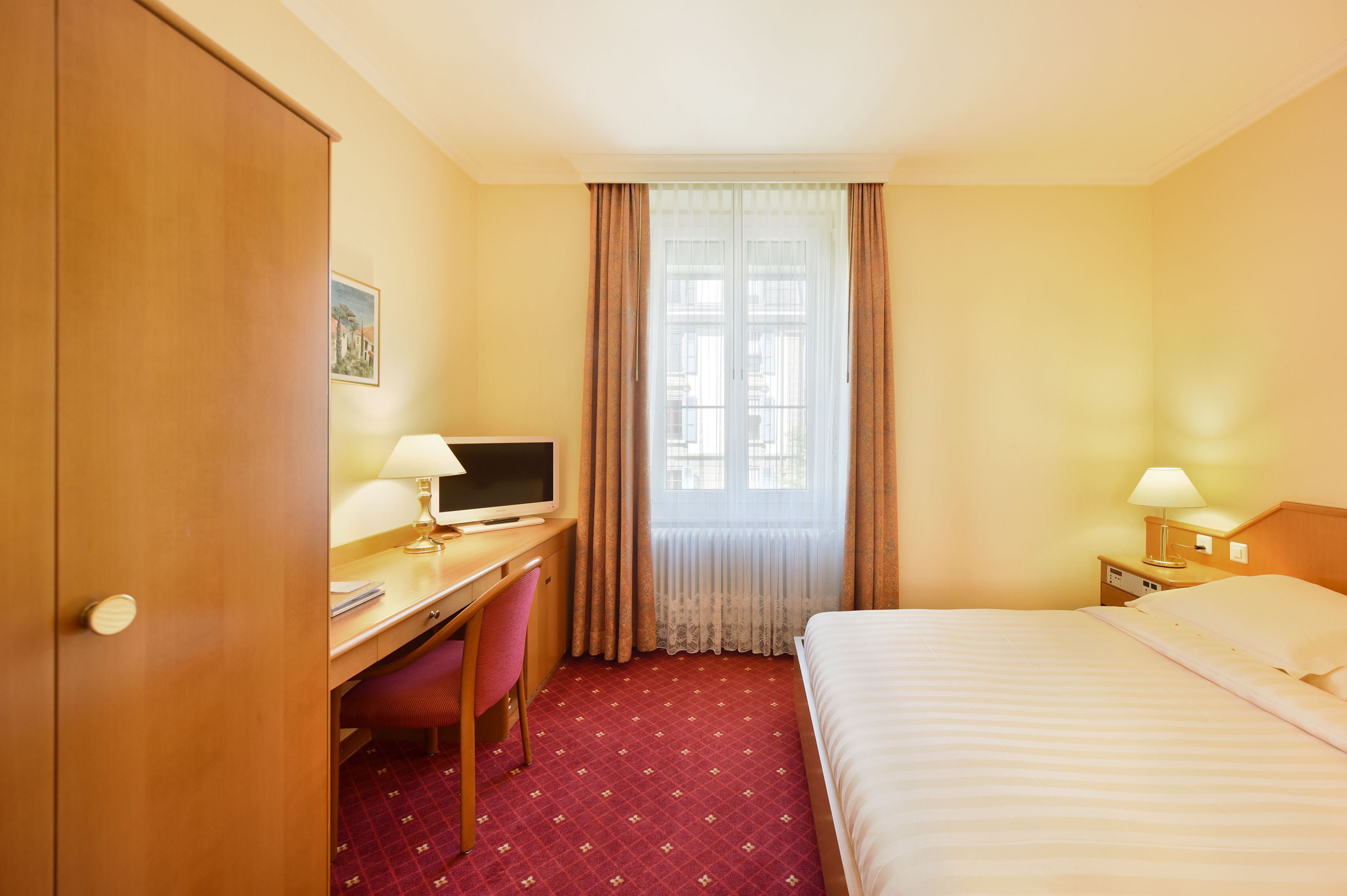Una habitación de hotel con cama, escritorio, silla y ventana con cortinas.