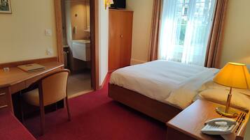 Habitación de hotel con cama, escritorio, silla y un baño visible a través de una puerta abierta.