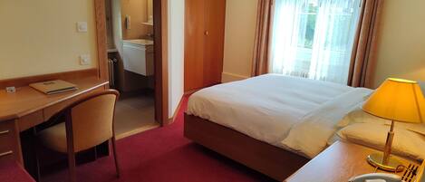 Quarto de hotel com cama, escrivaninha, cadeira e banheiro visível por uma porta aberta.