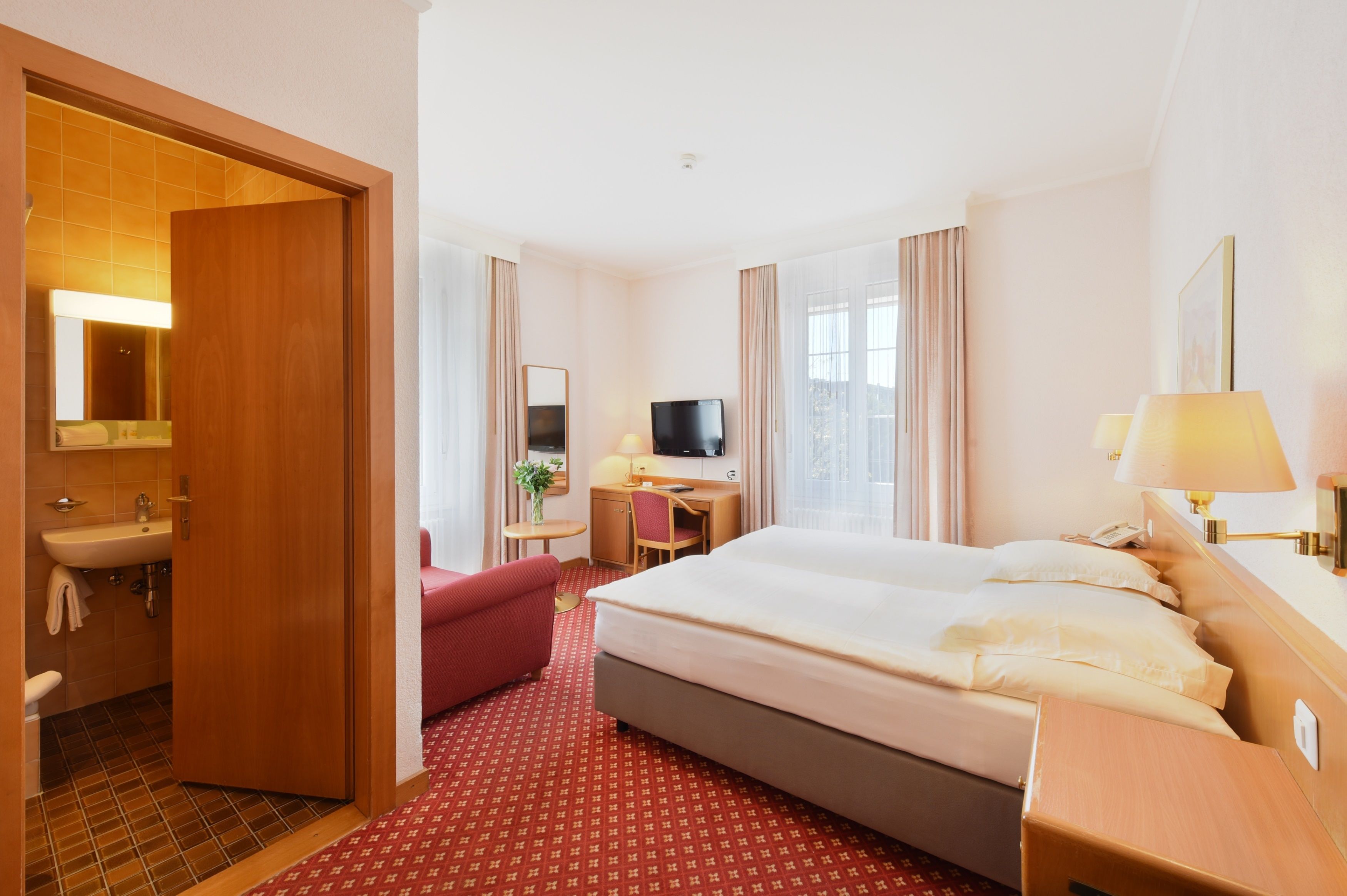 Una habitación de hotel con cama, escritorio, televisor y baño, además de una alfombra roja.
