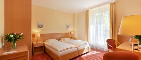 Habitación de hotel con cama doble, dos mesitas de noche con lámparas, un escritorio con silla, una mesita pequeña con un jarrón de flores y una ventana con cortinas.