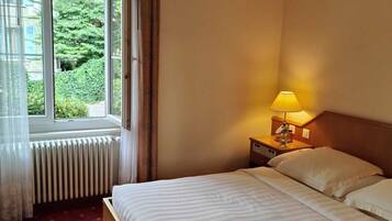 A hotel room with a bed, a nightstand with a lamp, a window with curtains, and a radiator.