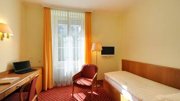 Ein Hotelzimmer mit Bett, Schreibtisch mit Laptop, Stuhl, Fernseher und Fenster mit Vorhängen.