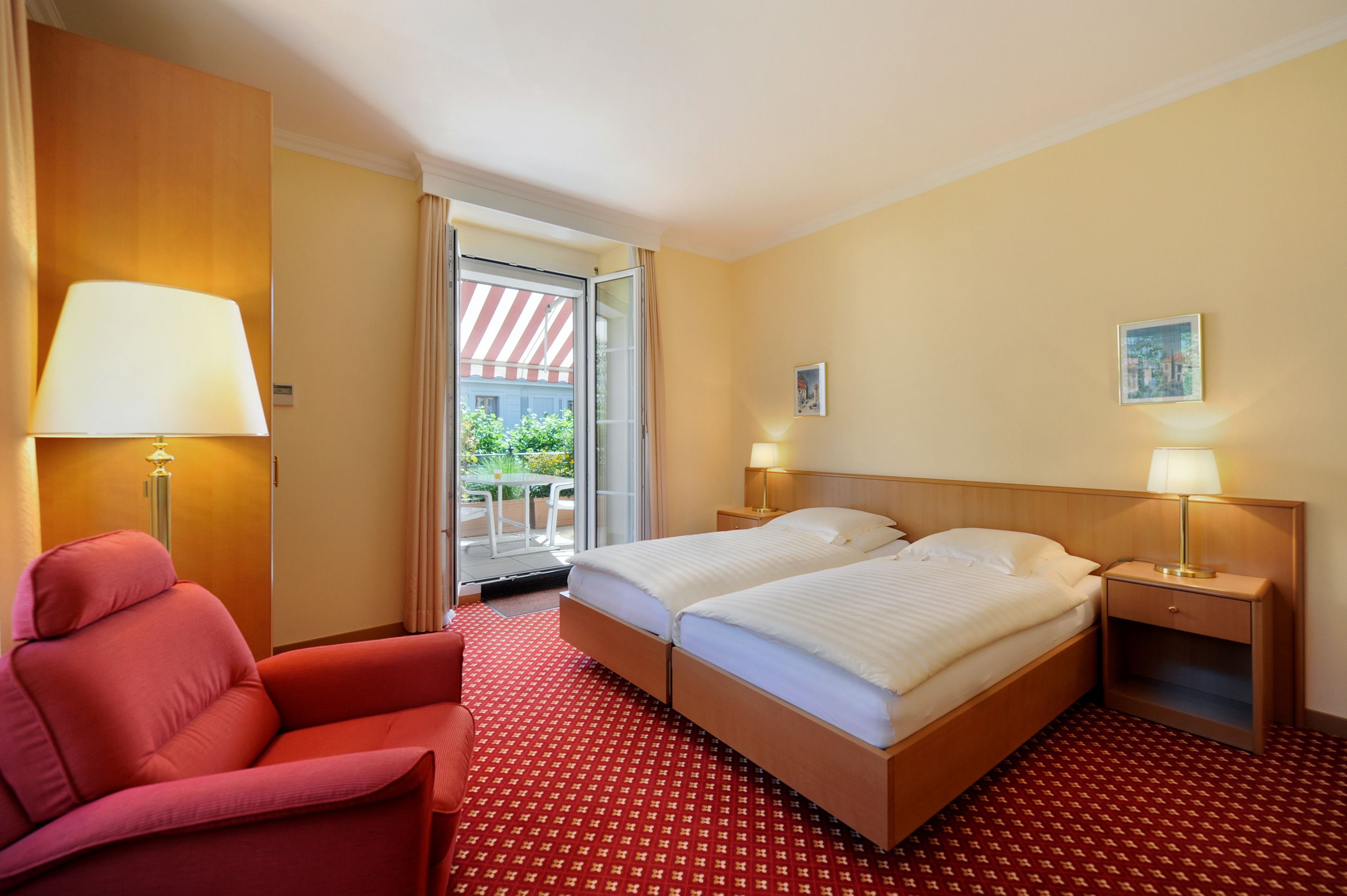 Habitación de hotel con dos camas, un sillón rojo, una mesita y vistas al exterior.