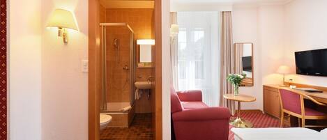 A hotel room with a shower, toilet, bed, desk, chair, TV, and a small table with a plant.