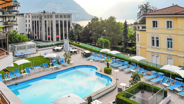 Zona de piscina con sillas de descanso y sombrillas, rodeada de edificios y un paisaje montañoso.