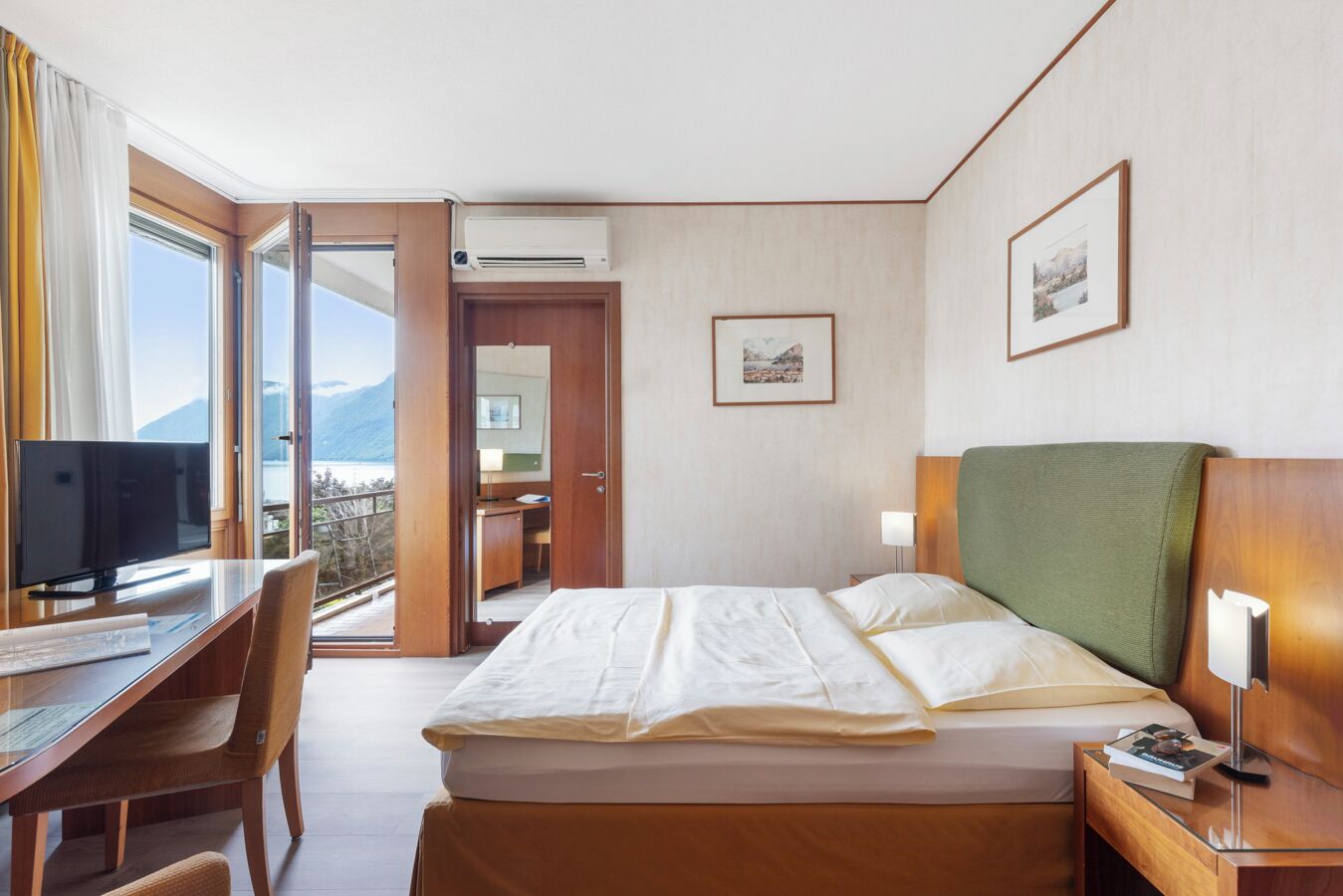 A hotel room with a bed, desk, and chair, a TV, and a view of a mountain and water.