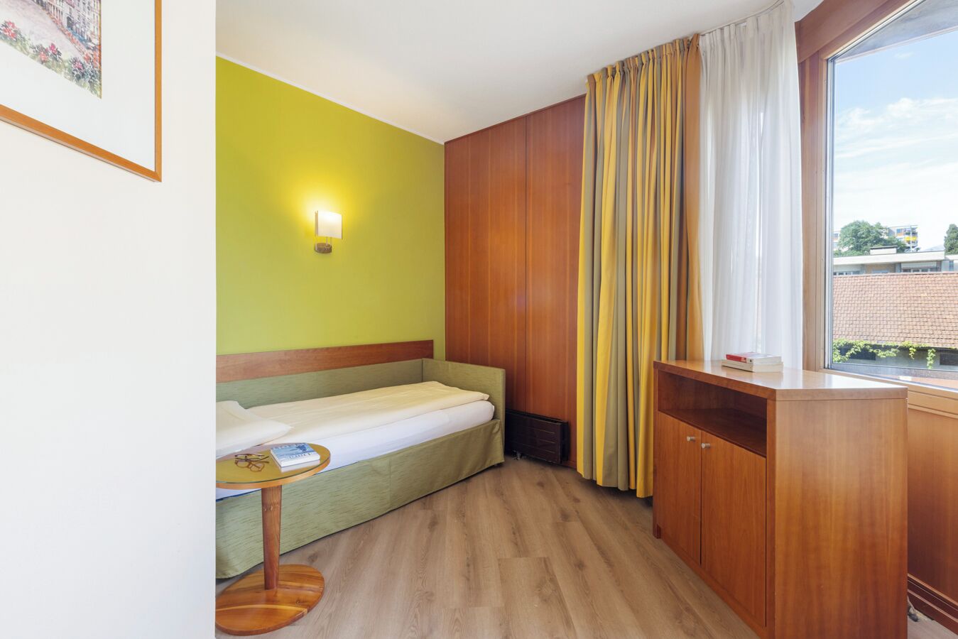 Habitación de hotel compacta con una cama individual, una mesita pequeña, un armario de madera y una ventana con cortinas.