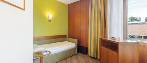 Habitación de hotel compacta con una cama individual, una mesita pequeña, un armario de madera y una ventana con cortinas.