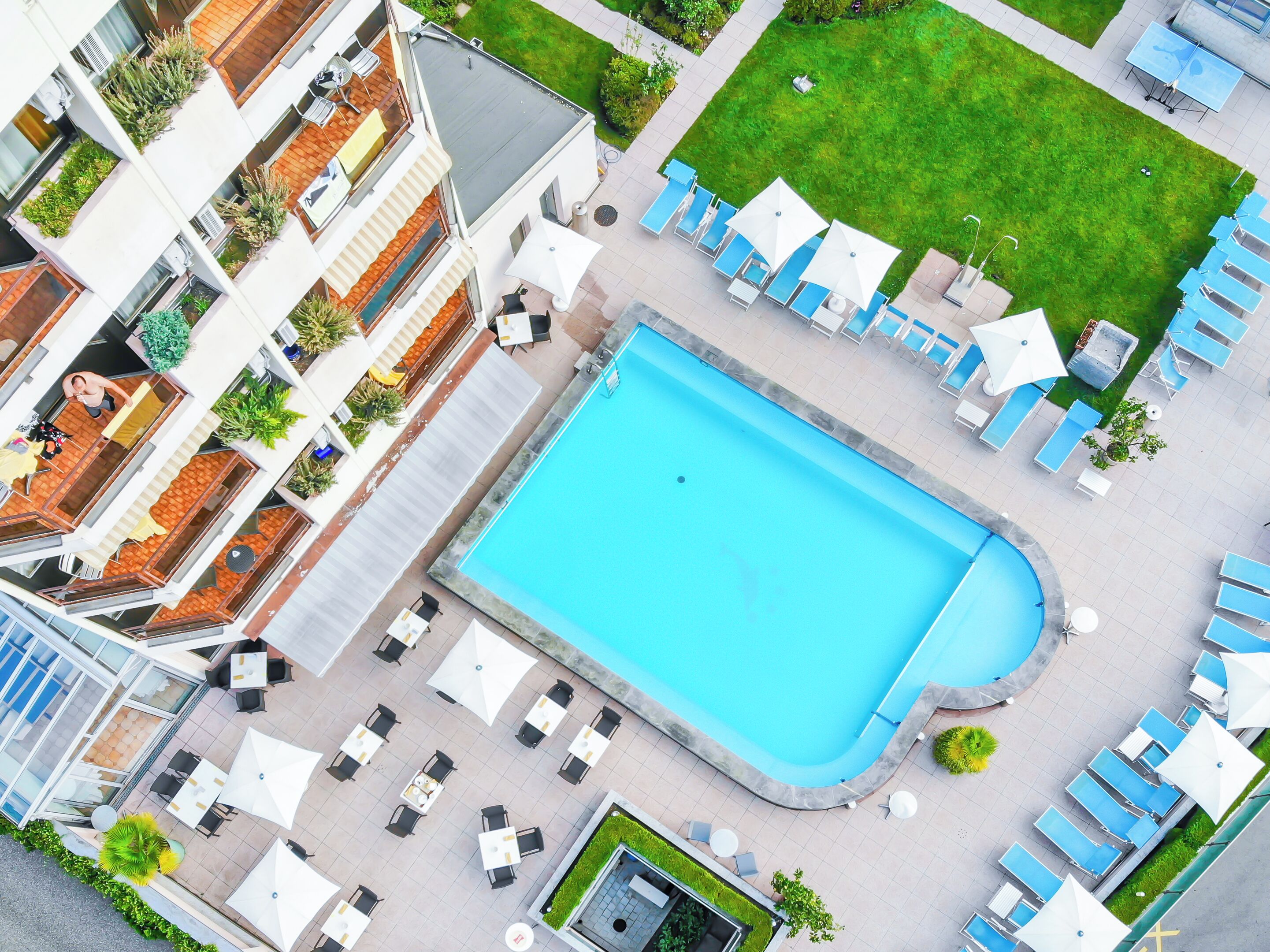 Ein Poolbereich auf dem Dach mit Liegestühlen und Sonnenschirmen, umgeben von einem Gebäude mit Balkonen und einer Rasenfläche.