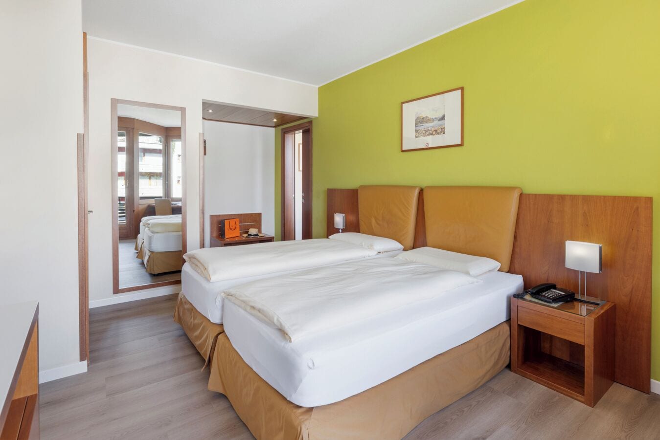 A hotel room with two beds, a wooden headboard, a bedside table with a lamp, and a framed picture on the wall.