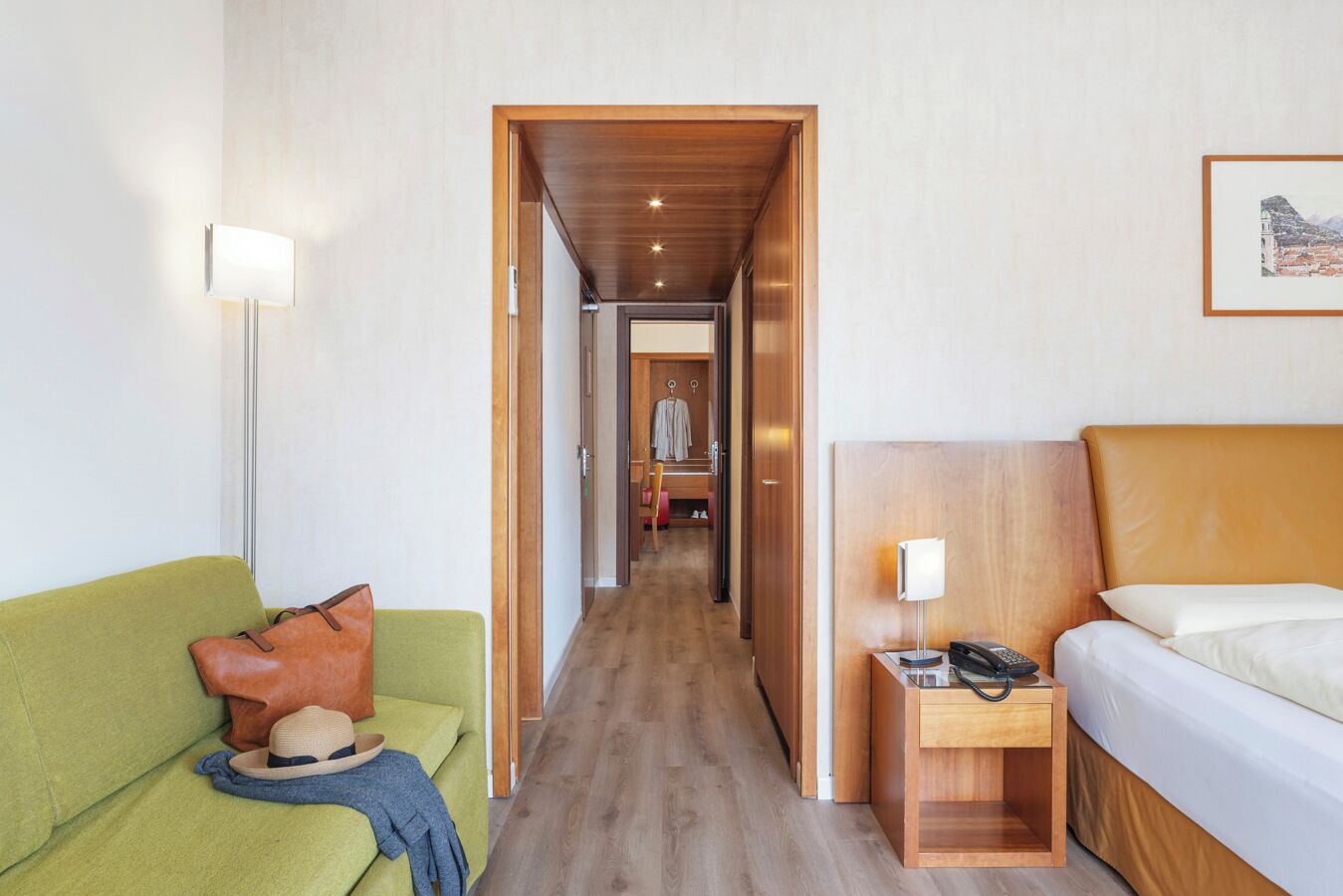 Een hotelkamer met een groene bank, een nachtkastje van hout met een lamp, een bed met hoofdeinde en een gang naar een badkamer.