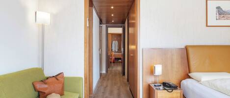 Habitación de hotel con un sofá verde, una mesita de noche de madera con una lámpara, una cama con cabecera y un pasillo que conduce a un baño.