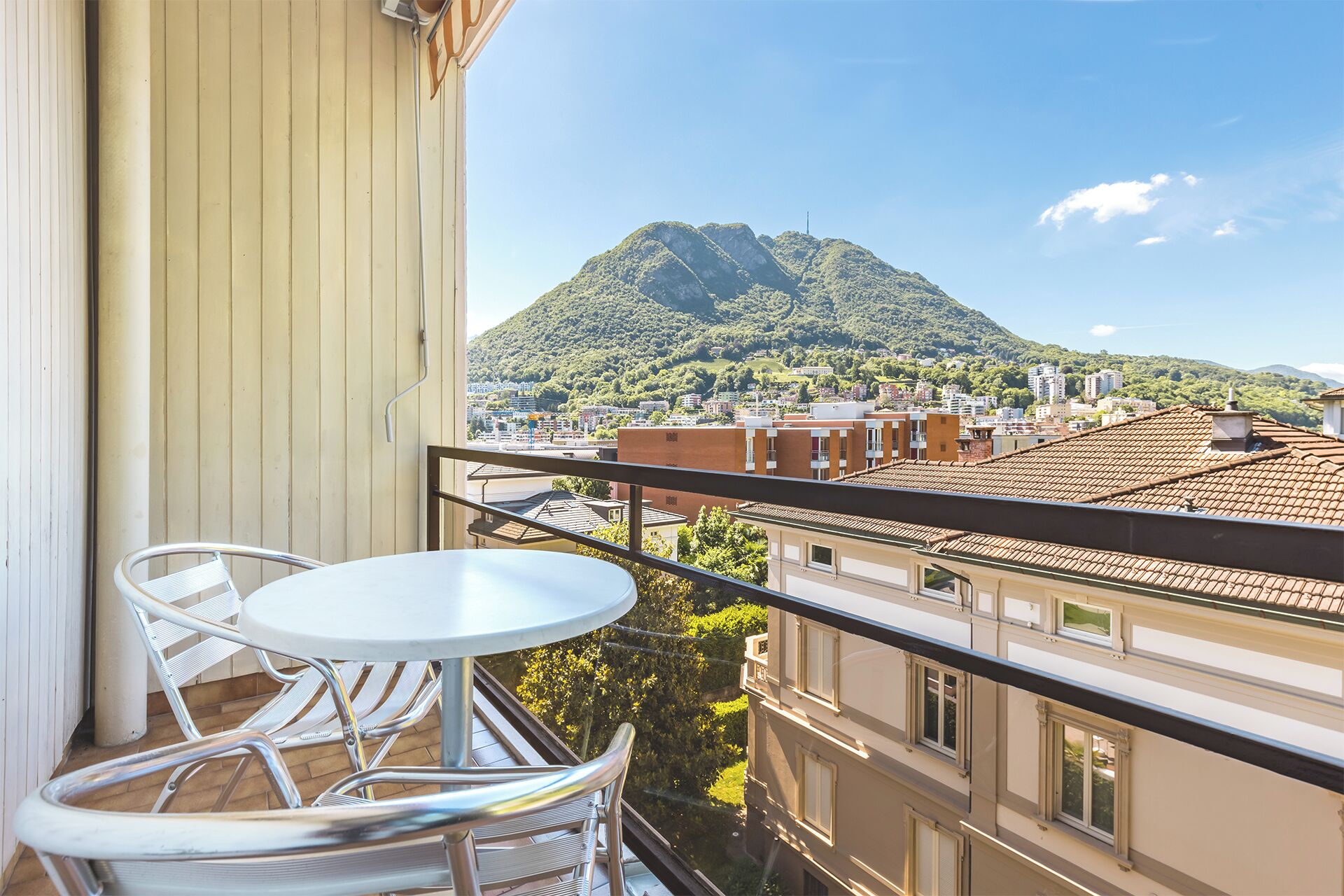 A balcony with a table and chairs, overlooking a cityscape and a mountain.