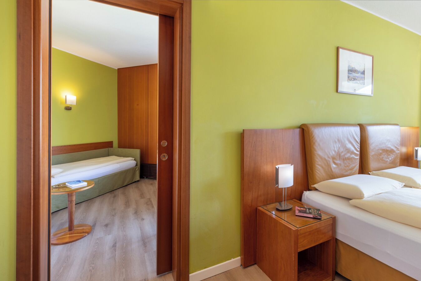 A hotel room with a bed, a bedside table with a lamp, a small table with a book, and a door leading to another room.