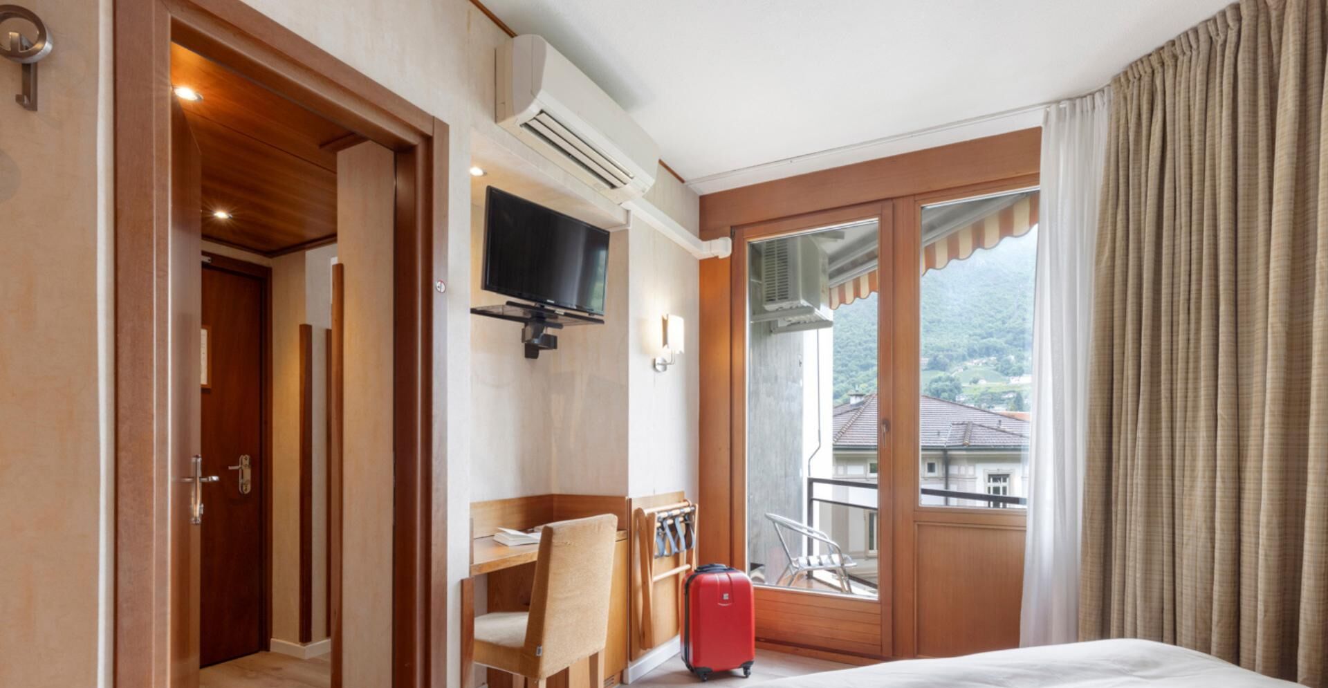 Camera d'albergo con un letto, una scrivania, una sedia, una TV e un balcone con tende.