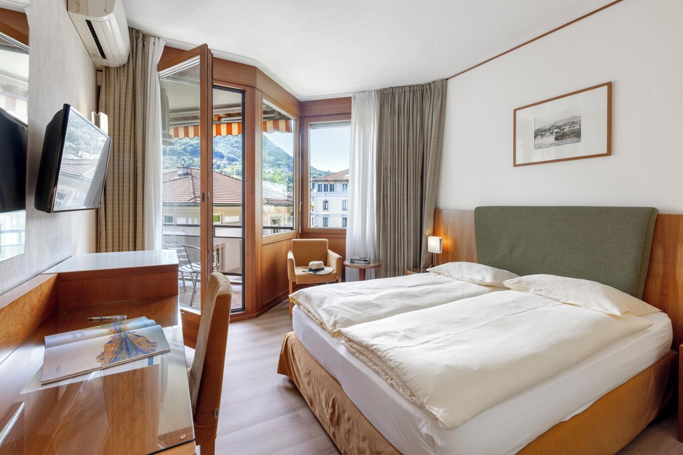 A hotel room with a bed, a desk, a chair, a TV, and a balcony with curtains.