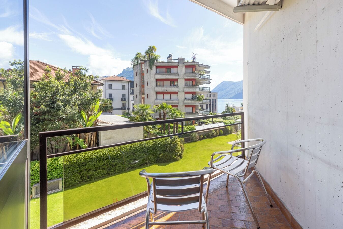 Un balcón con dos sillas y vista a un jardín verde, con un edificio que tiene balcones al fondo.