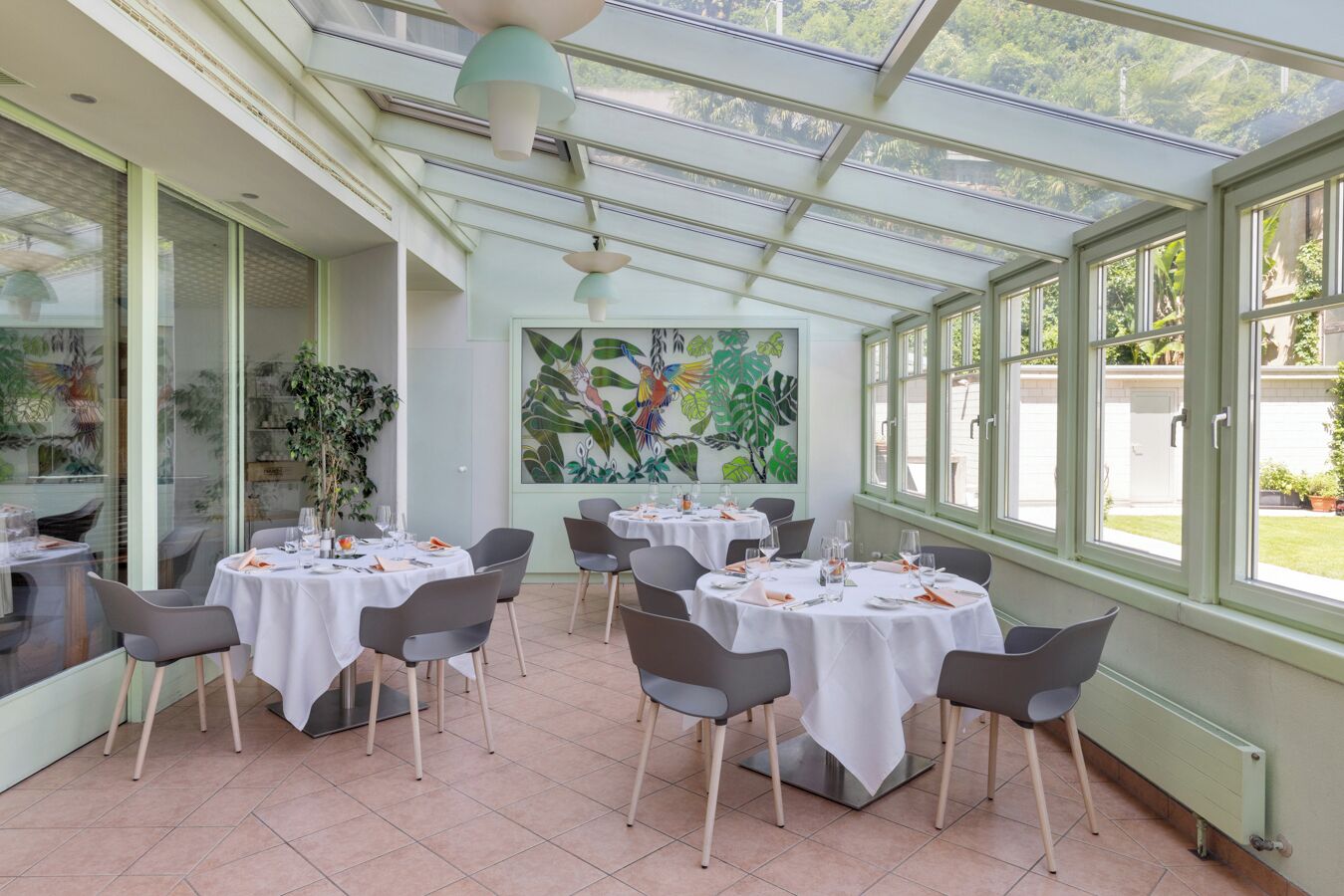 A restaurant with round tables, grey chairs, and a large mural of birds and foliage.