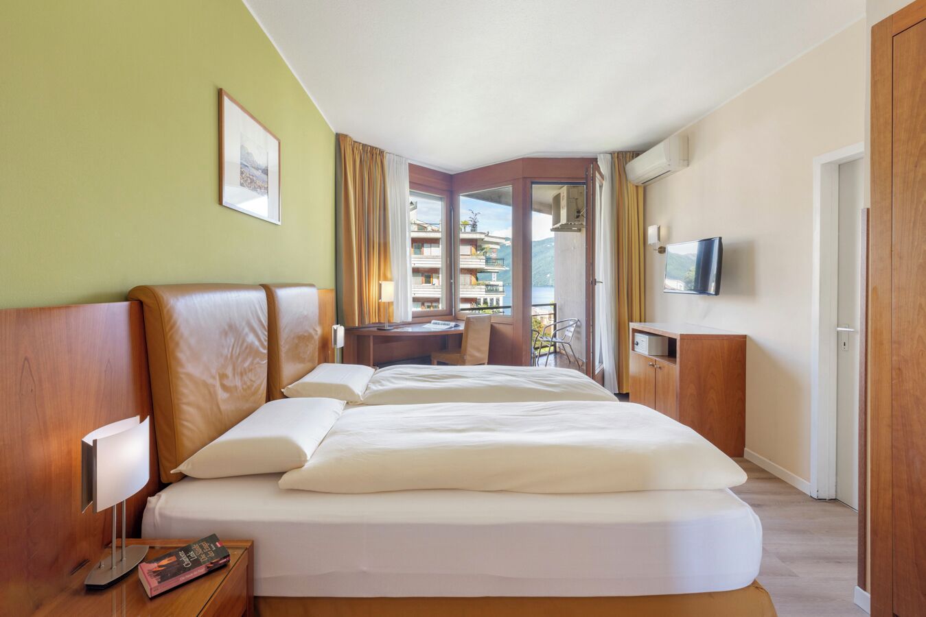 Une chambre d’hôtel avec un grand lit, un bureau, une chaise, une télévision et un balcon donnant sur les bâtiments.