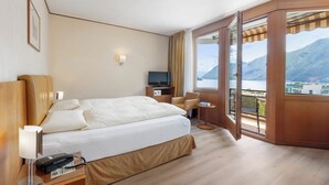 Habitación de hotel con una cama grande, un escritorio con silla, televisor y vistas a un lago y montañas.