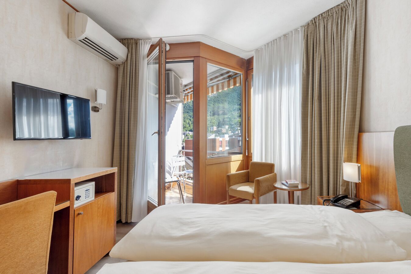 Quarto de hotel com cama, escrivaninha, cadeira, televisão e janela com cortinas.