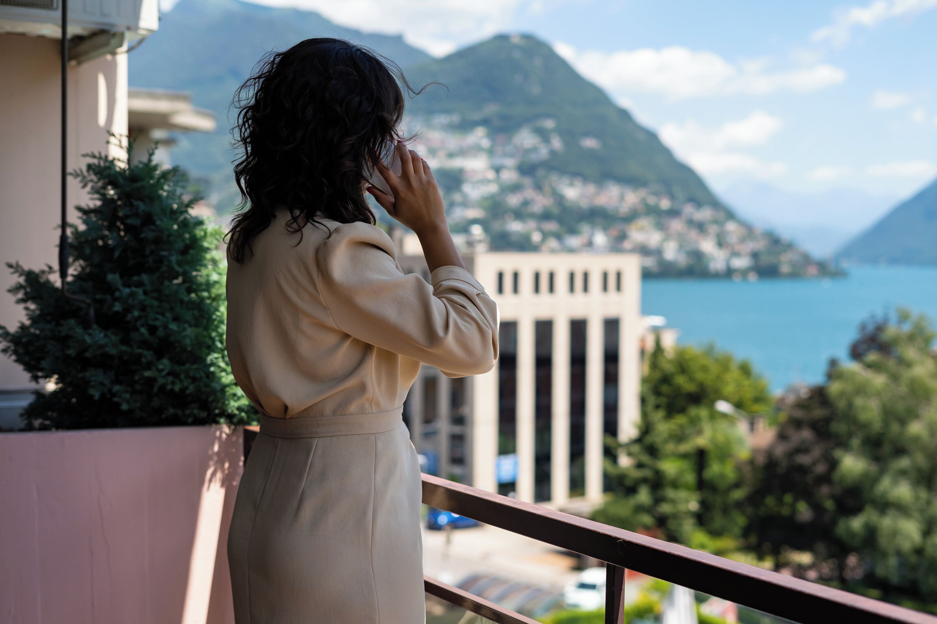 Eine Person steht auf einem Balkon mit Blick auf eine gebirgige Landschaft, in der sich ein Gewässer und Gebäude in der Ferne befinden.