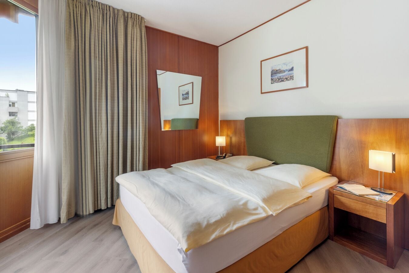 A hotel room with a bed, two bedside tables, a window with curtains, a wall-mounted picture, and a view of the outside.