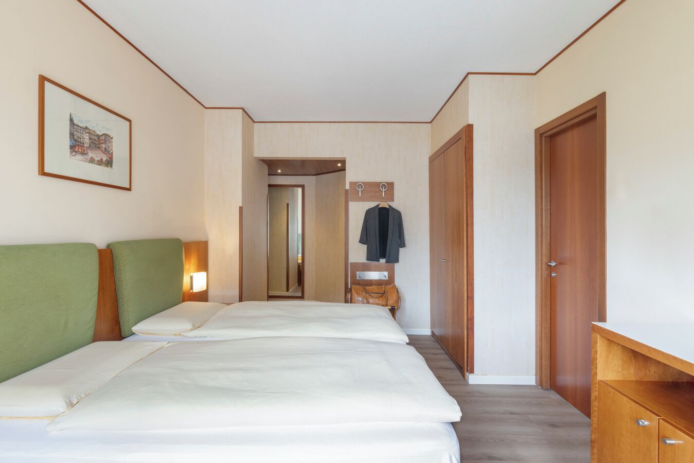 A hotel room with two beds, a wooden wardrobe, a coat rack, and a framed picture on the wall.
