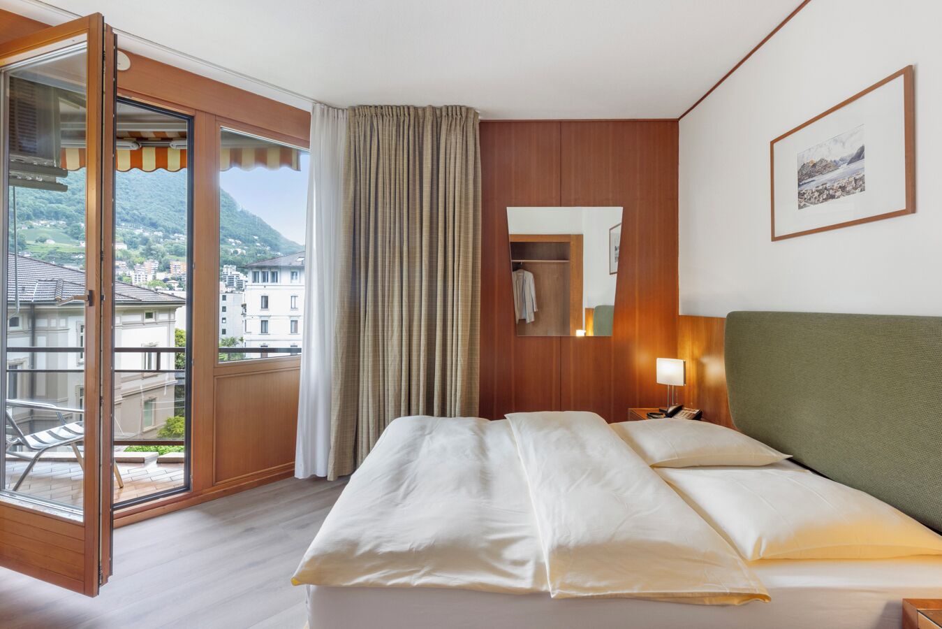 A hotel room with a bed, a bedside table with a lamp, a mirror, and a view of a balcony with a cityscape.