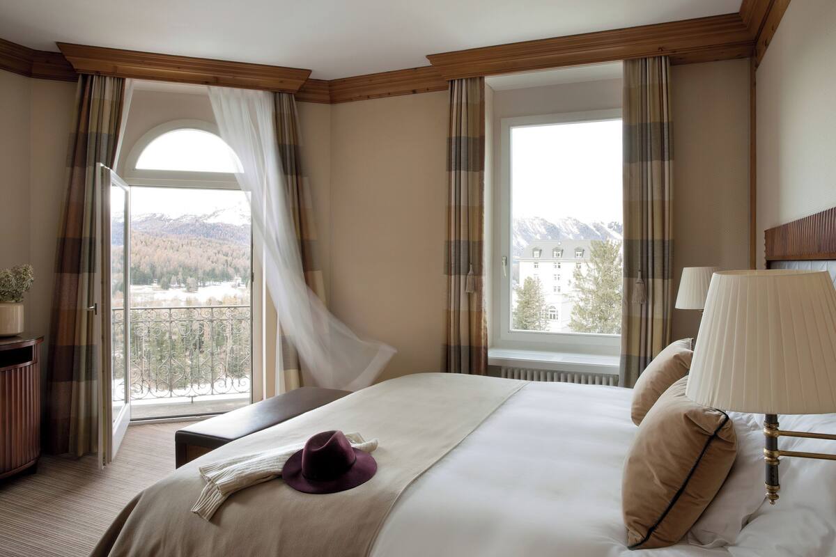 A hotel room with a large bed, a bedside table, a lamp, and a view of mountains through the window.