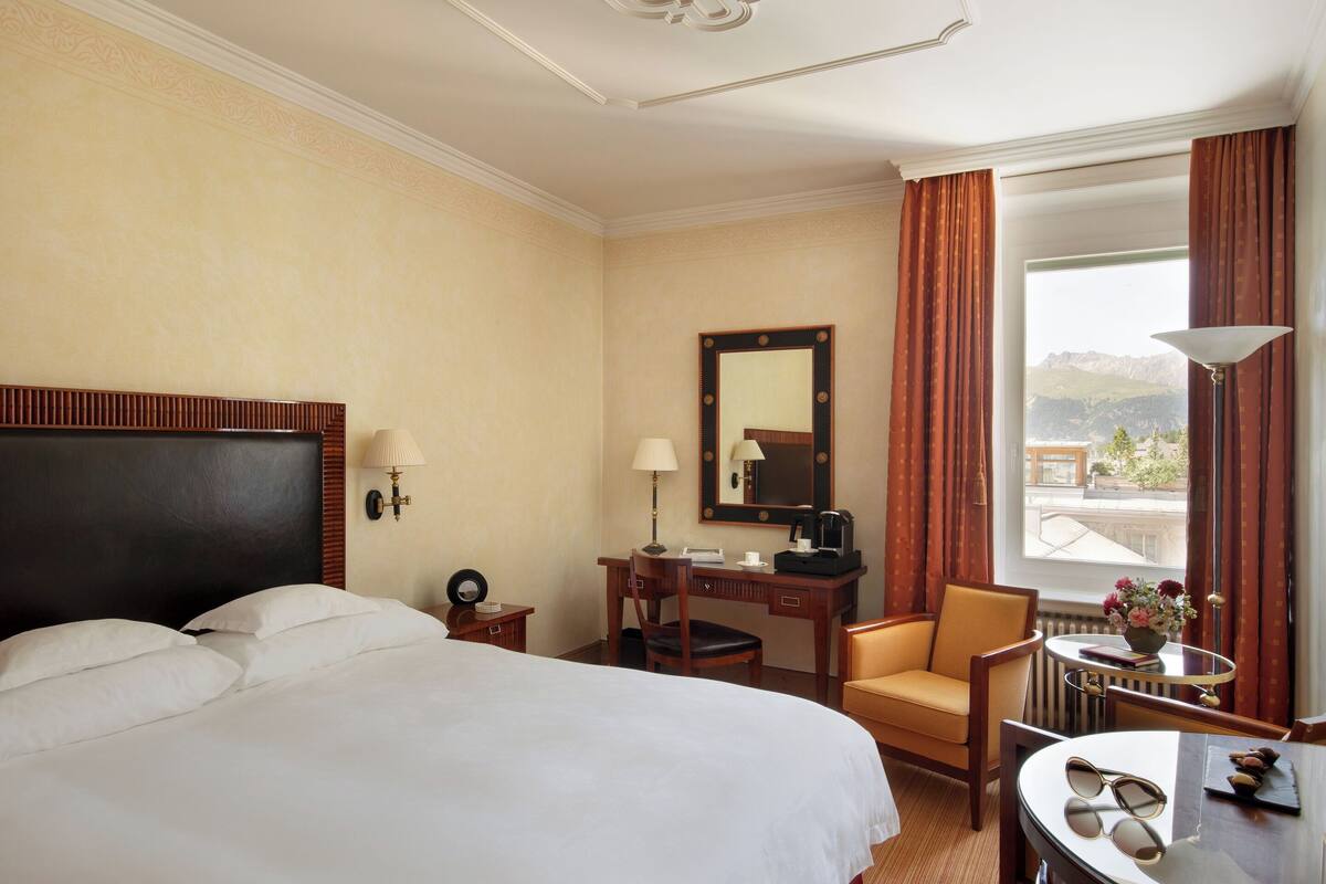 A hotel room with a large bed, a desk with a chair, a small table with a lamp, and a view of mountains through the window.