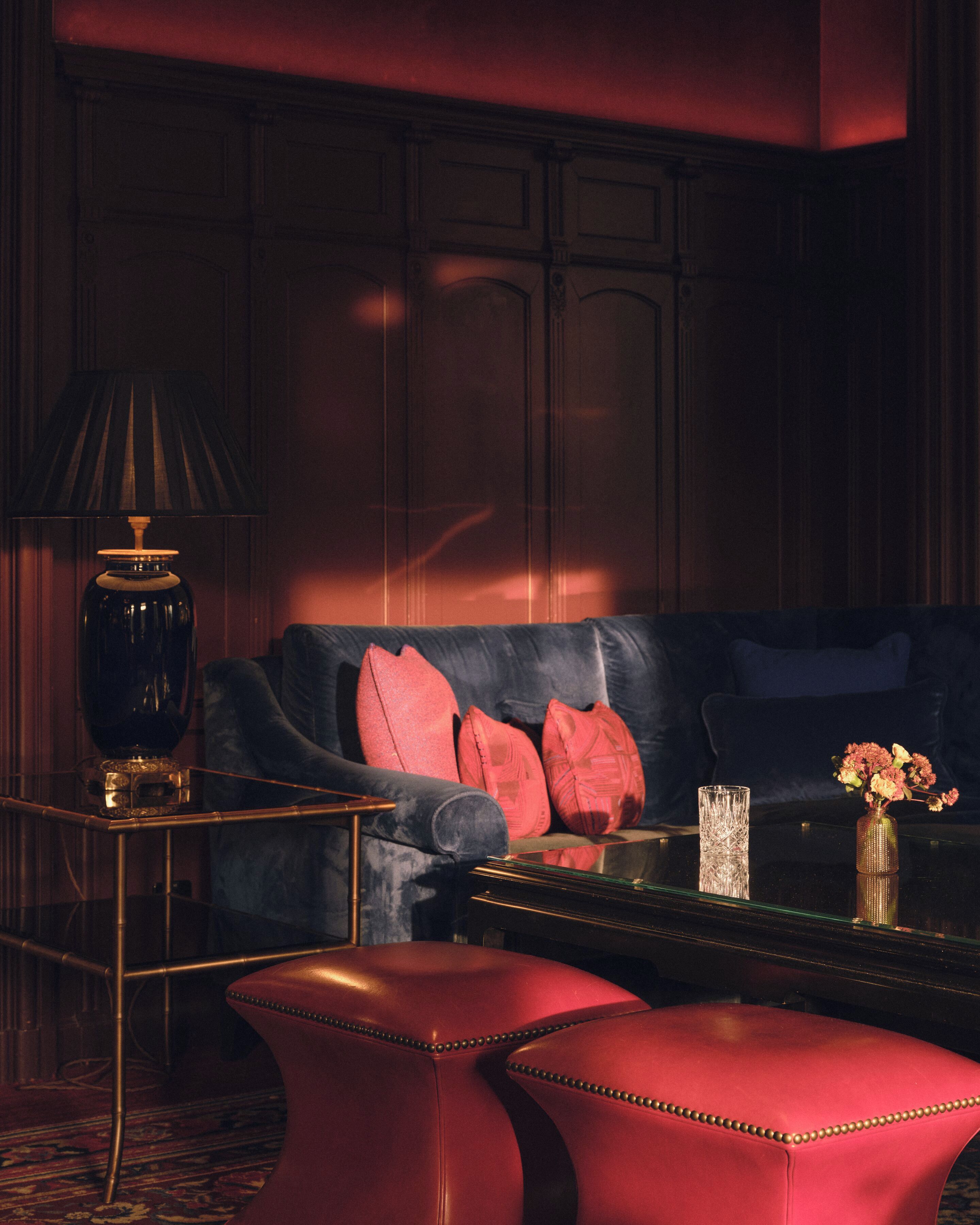 Un salón con decoración oscura, un sofá de cuero, cojines rojos, una mesa de centro de vidrio y una lámpara.