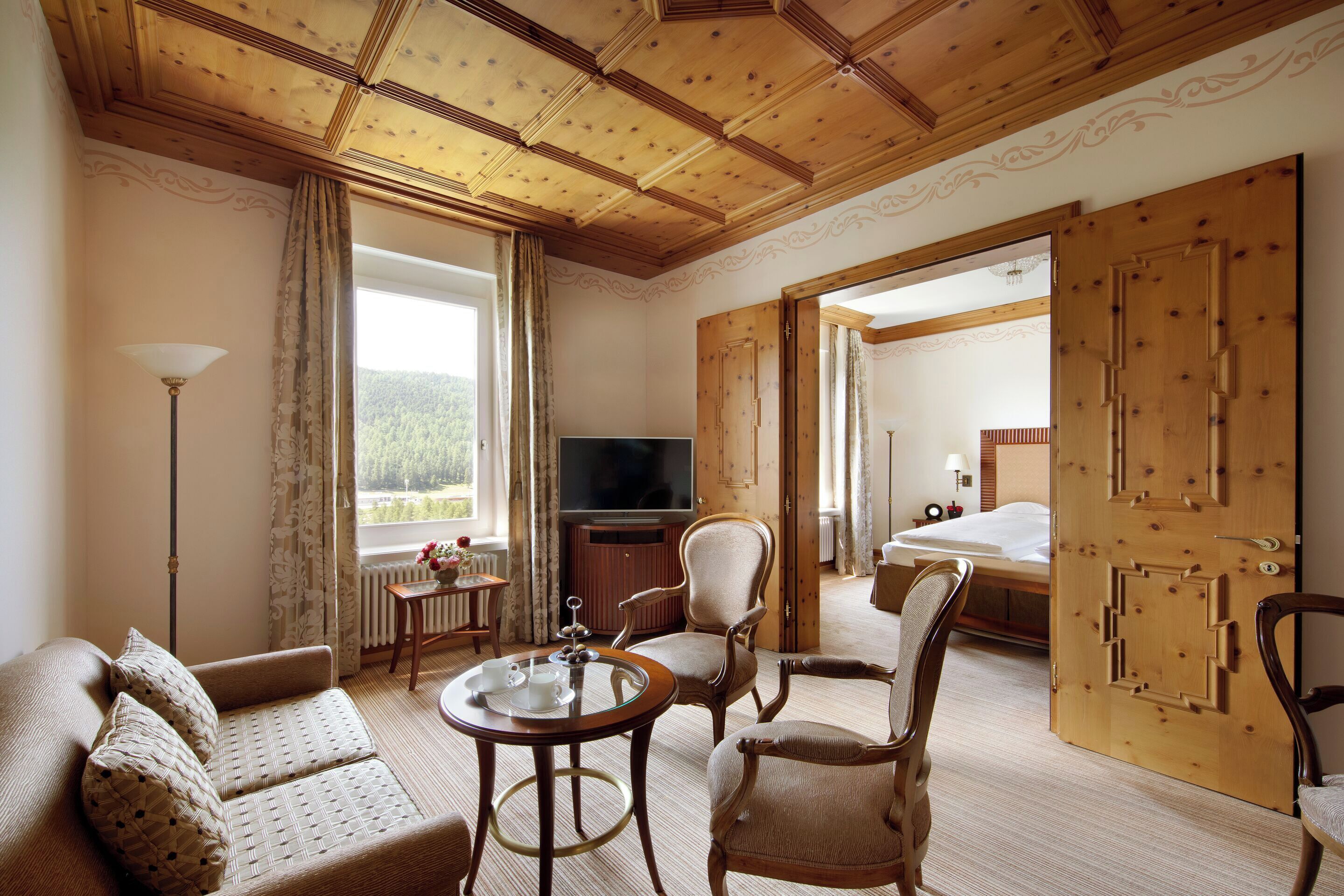Habitación de hotel con techo de madera, un sofá, dos sillones, un televisor, una mesita con bandeja y una puerta grande de madera.