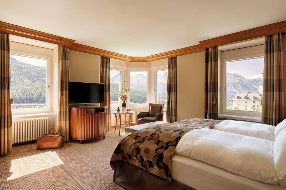 A hotel room with a large bed, a television, a small table with a plant, and a view of mountains through large windows.