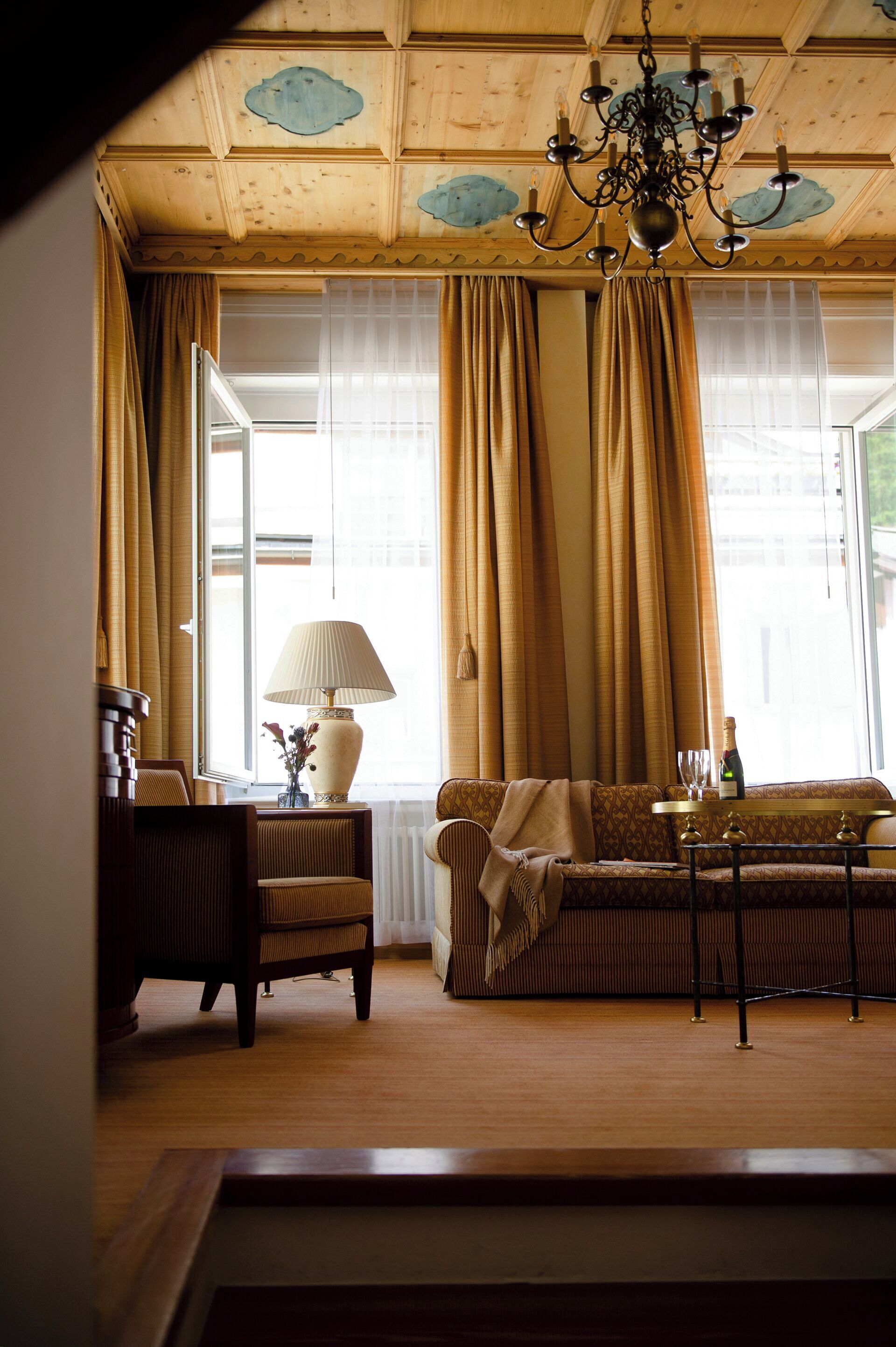 Una habitación acogedora con techo de madera, una araña, un sofá, una mesa con lámpara y una ventana con cortinas.