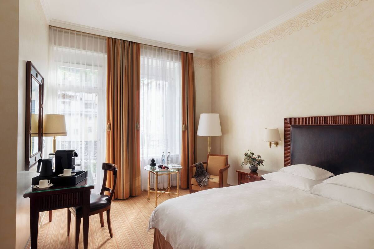 A hotel room with a bed, a desk with a coffee maker, a chair, a small table with a lamp, and a window with curtains.