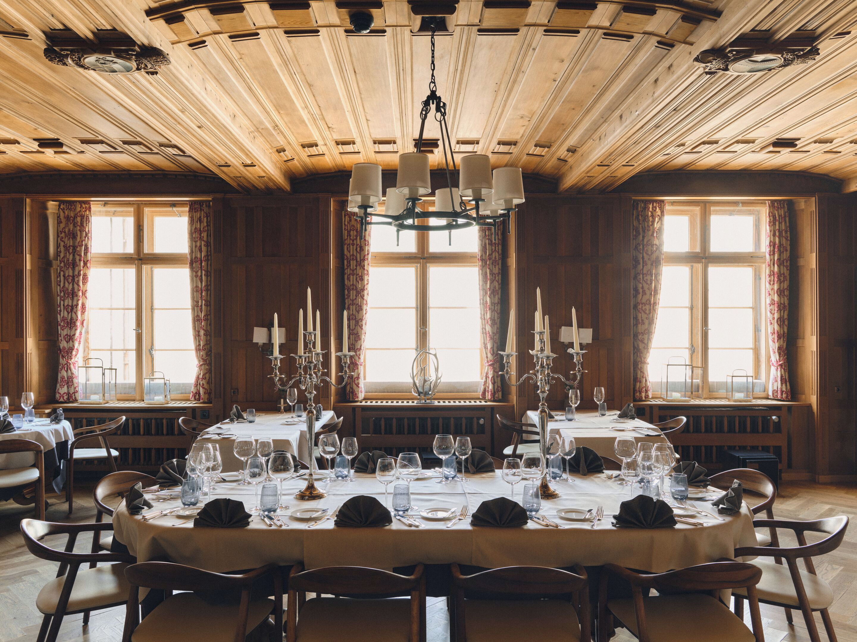 Une salle à manger avec une grande table en bois dressée pour un repas, flanquée de hautes fenêtres ornées de rideaux, et un plafond en bois avec des poutres apparentes.