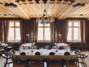 A dining room with a large wooden table set for a meal, flanked by tall windows with curtains, and a wooden ceiling with exposed beams. - Grand Hotel Kronenhof (Pontresina)