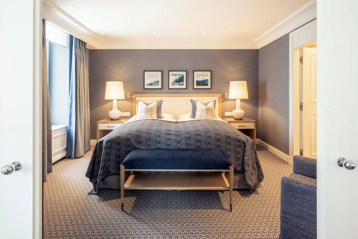 A hotel room with a large bed, two bedside tables with lamps, a bench, and a view of the outside.