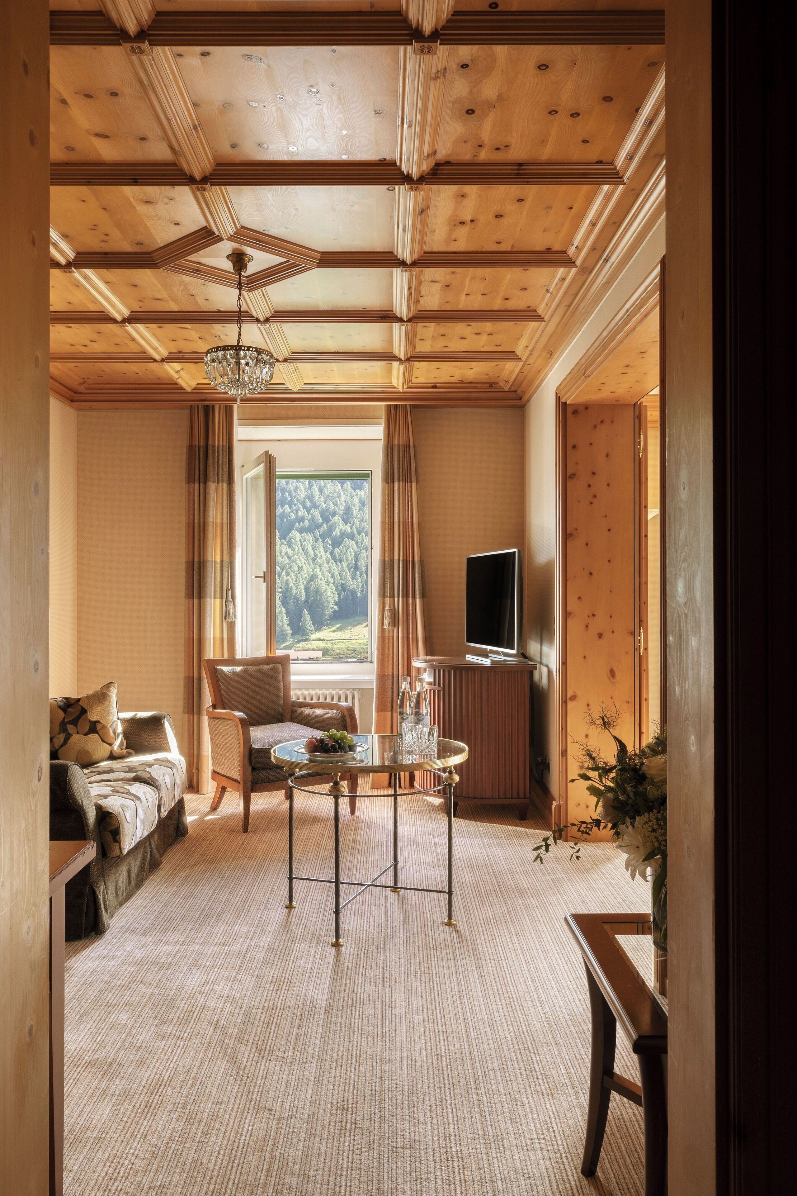 Un salon chaleureux avec un plafond en bois, un canapé, un fauteuil, une table ornée d’un vase et un téléviseur posé sur un meuble.